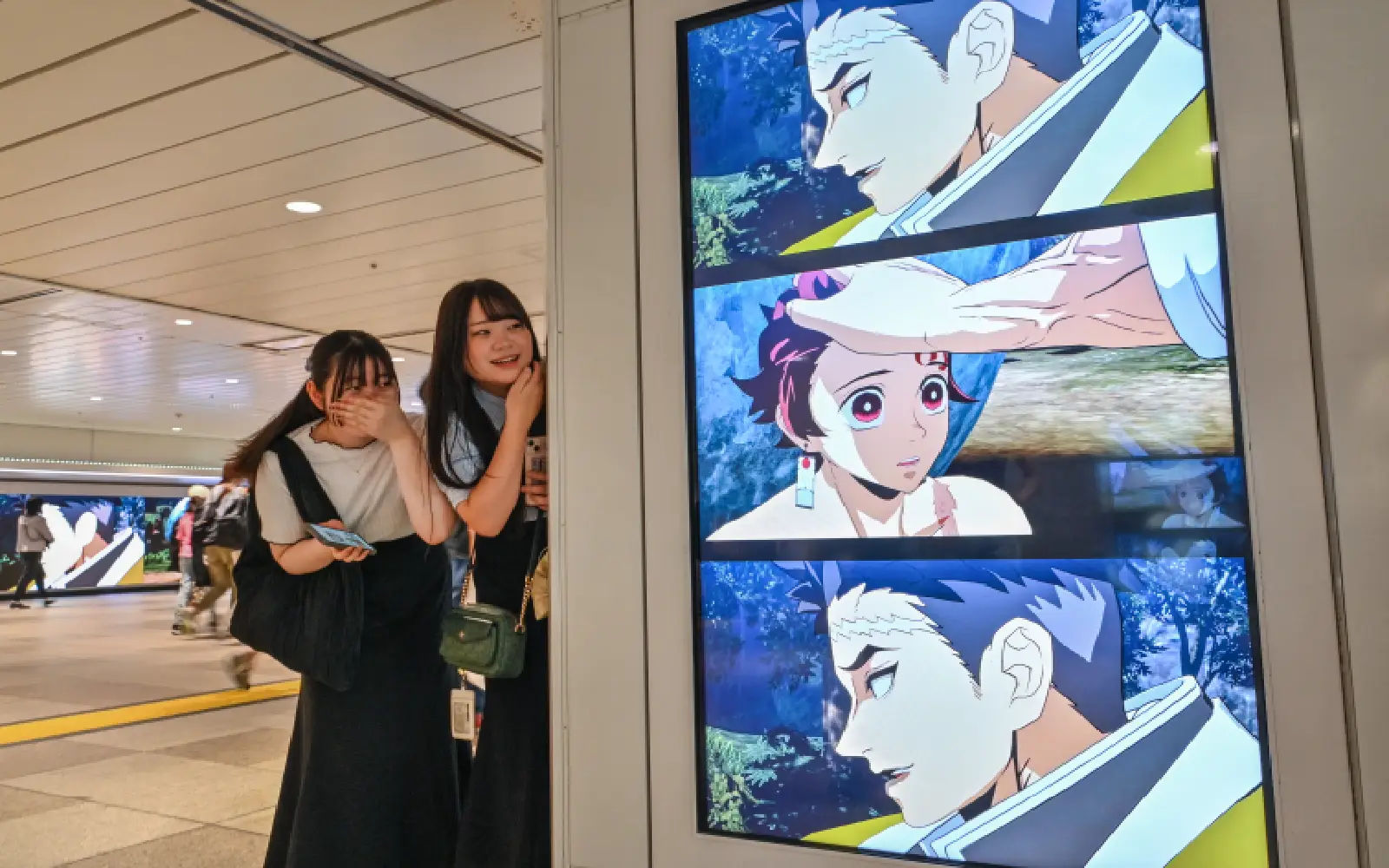 Two young women look over at a friend as they stand next to a digital billboard advertising the upcoming release of the video game "Demon Slayer: Kimetsu no Yaiba – The Hinokami Chronicles 2", along a walkway inside a train station in central Tokyo on July 29, 2025. As a teenager struggling to fit into life in rural Sicily, psychiatrist Francesco Panto found refuge in anime, where he discovered characters that resembled the kind of man he wanted to be.